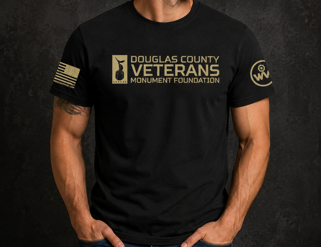 A man wearing a black short-sleeve t-shirt with a gold-colored 'Douglas County Veterans Monument Foundation' logo on the chest, a gold American flag graphic on the sleeve, and a circular 'SW' logo on the other sleeve, set against a dark textured background.