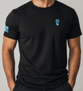 A man wearing a black short-sleeve crewneck t-shirt with a small light blue logo on the left chest and a light blue graphic on the right sleeve, photographed against a neutral gray studio background.