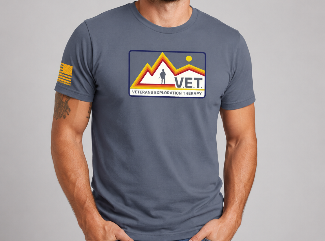 A man wearing a slate blue-gray t-shirt with a 'VET VETERANS TRUST' graphic on the chest and a US flag patch on the sleeve, paired with blue jeans, against a solid gray studio background.