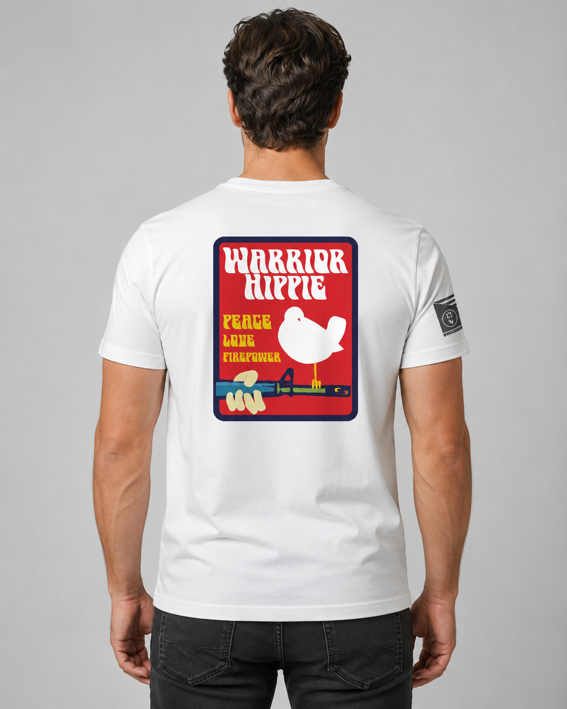 A man viewed from behind wearing a white t-shirt with a large red, yellow, and blue graphic on the back that reads 'WARRIOR HIPPIE PEACE AND LOVE' with a white bird illustration, and a small black square patch on the left sleeve.